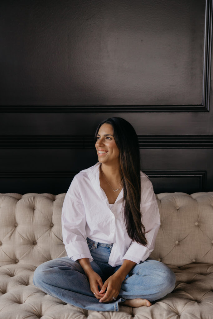 Tatiana Budney in a white button-down and jeans, seated on a tufted couch, looking up and smiling