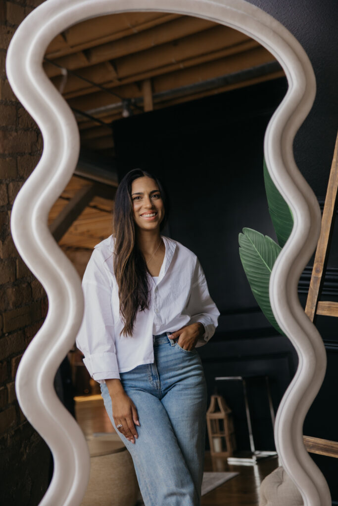 Tatiana Budney in a white button-down and jeans, standing beside a wavy mirror in a loft space