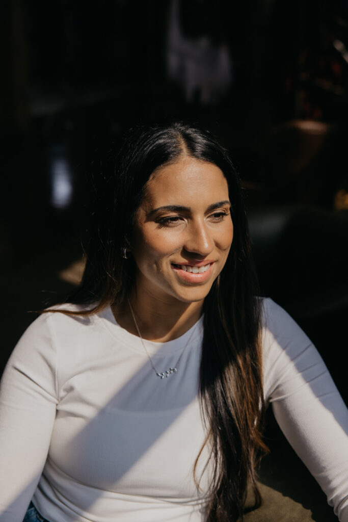 Tatiana Budney in a white long-sleeve top, sunlight across her face, smiling in a loft studio