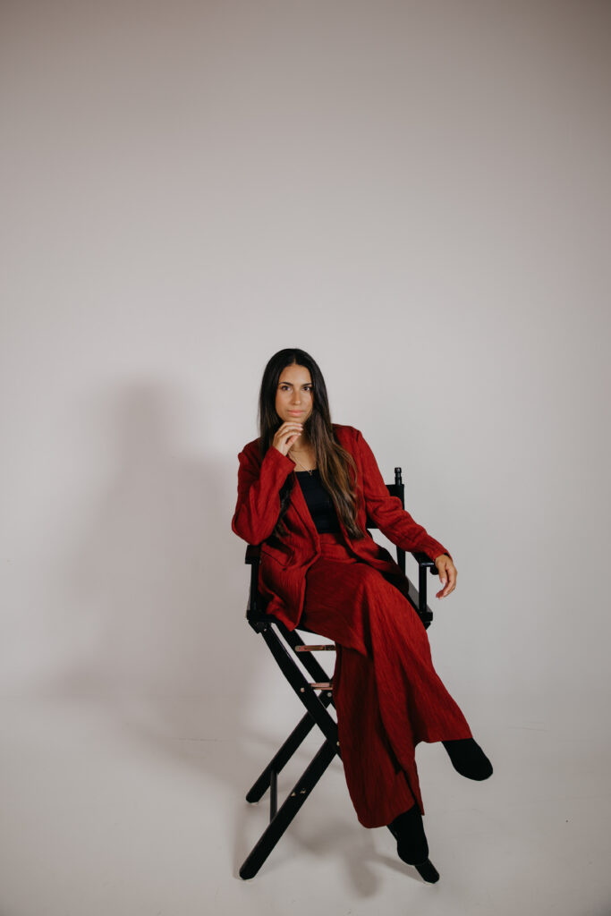 Tatiana Budney seated in a red duster suit, hand near chin, thoughtful smile on set