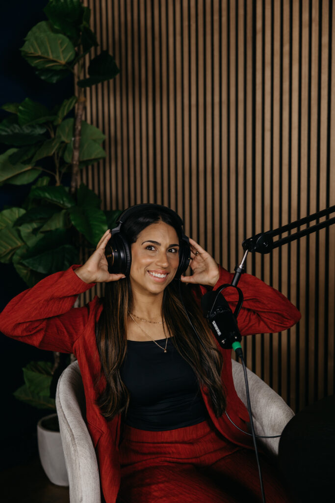 Tatiana Budney in a red duster suit, hands on headphones, laughing in a podcast studio