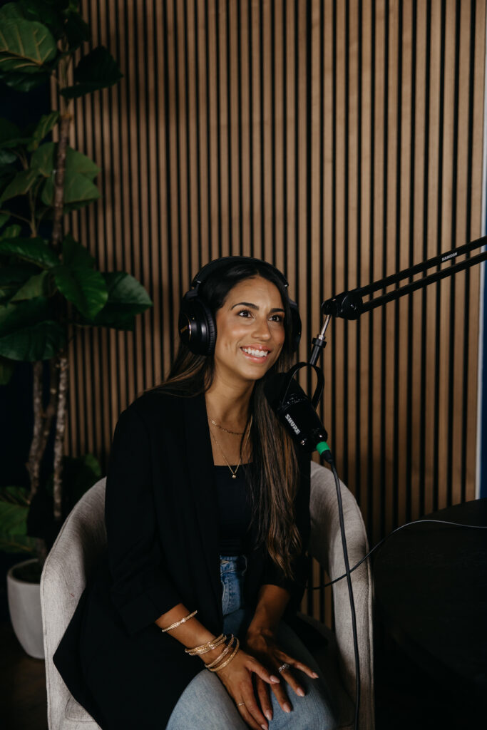 Tatiana Budney in a black blazer and headphones, smiling in a podcast studio with wood slat wall