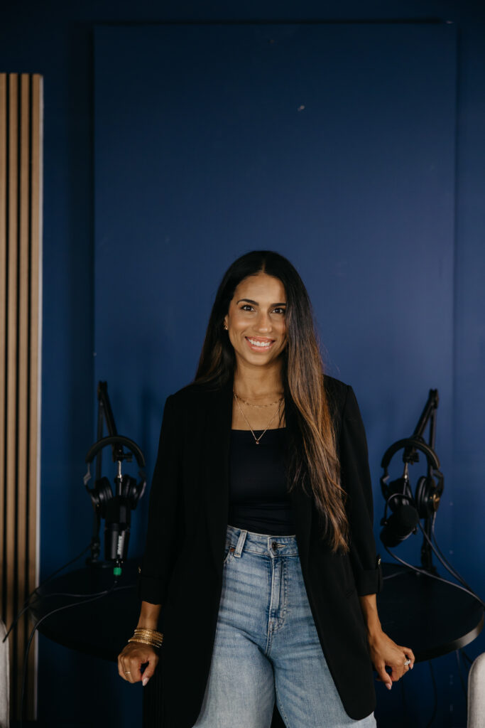Tatiana Budney in a black blazer and jeans standing in a podcast studio with mic and headphones