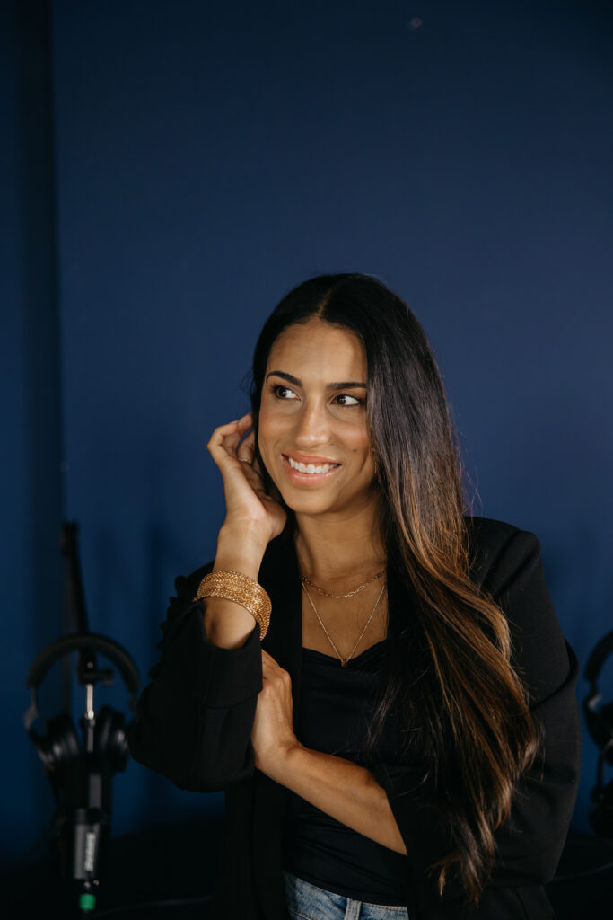 Tatiana Budney in a black blazer against a navy backdrop, hand near ear, studio mic visible