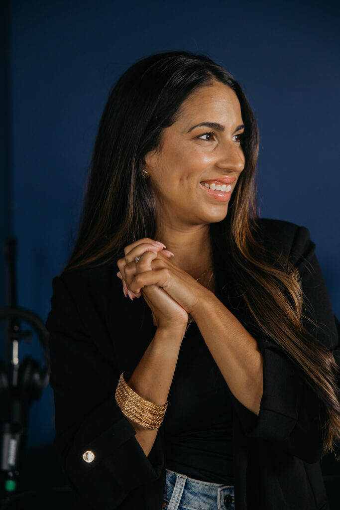 Tatiana Budney in a black blazer, hands clasped, smiling against a navy studio backdrop