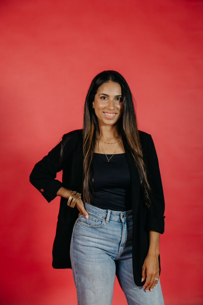 Tatiana Budney in a black blazer and jeans, hand on hip, against a bold red backdrop