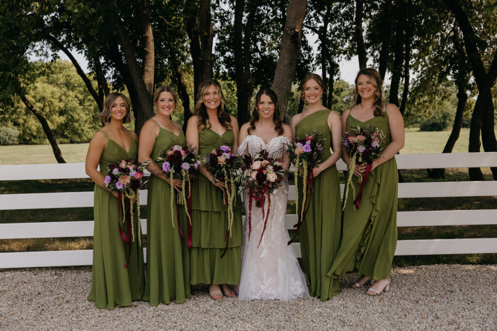 Bridesmaids in olive green dresses standing in front of a white fence