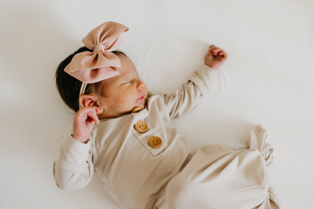 Newborn baby girl in a cream buttoned outfit with a pink bow headband, lying on a white blanket with eyes closed