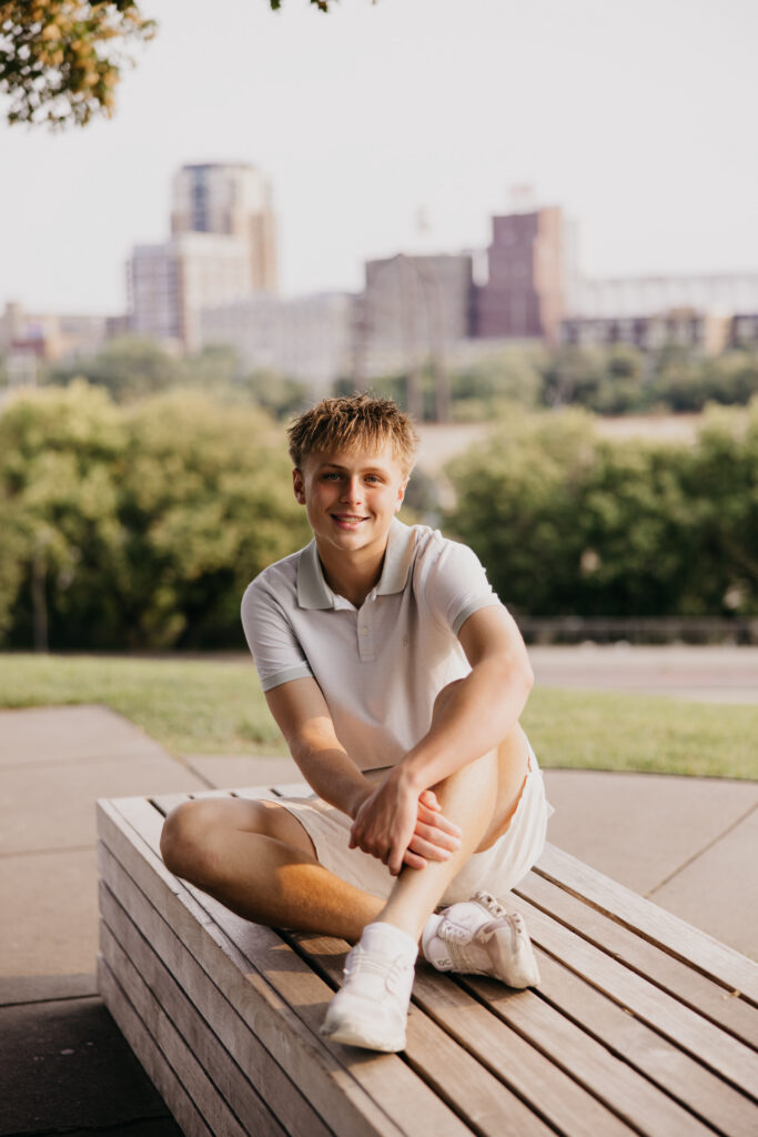 Senior guy in a gray polo sitting on a wooden bench with the Minneapolis skyline behind him