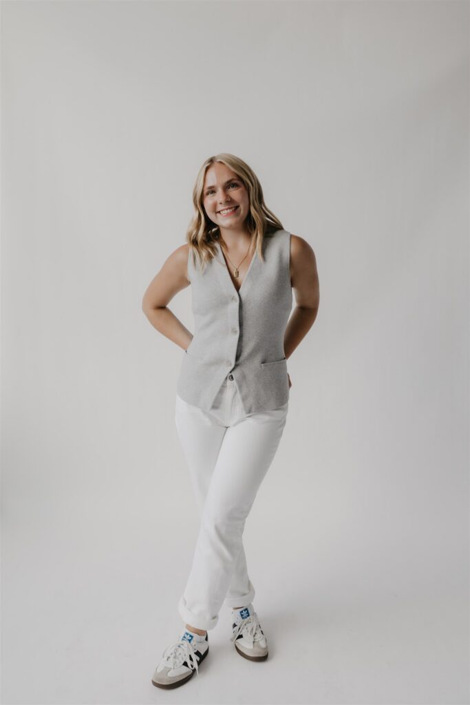 Woman in a gray tailored vest, white pants, and white sneakers smiling in a studio