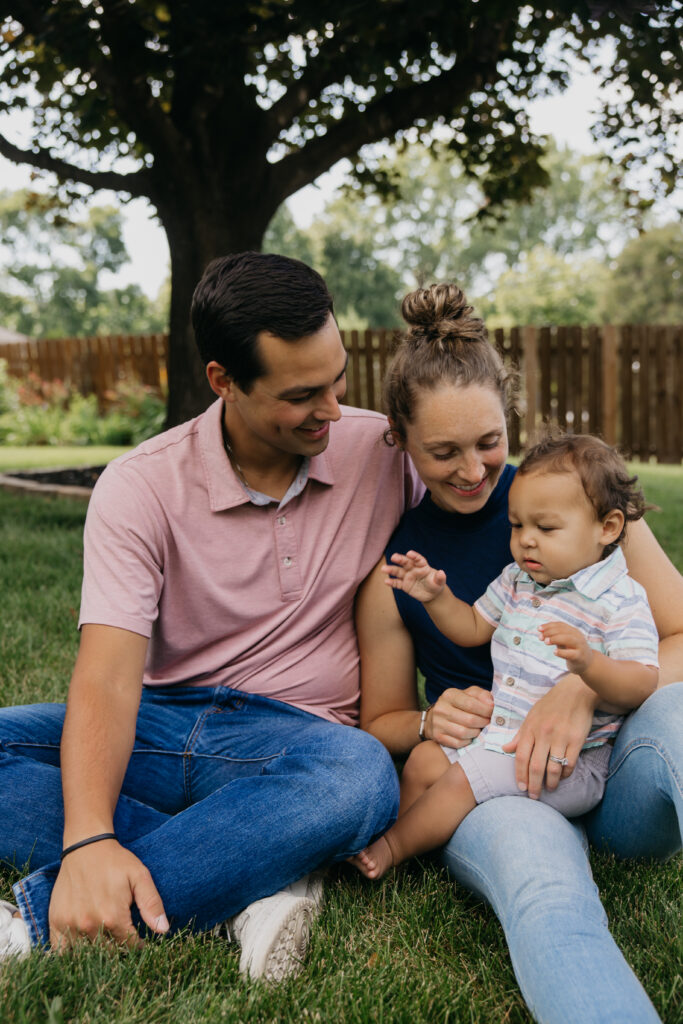 Mom and dad sitting on the grass under a tree with baby between them, backyard family photos in Minneapolis
