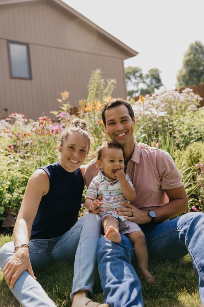 Mom and dad sitting in a garden with baby on dad's lap, blooming flowers and a shed in the background
