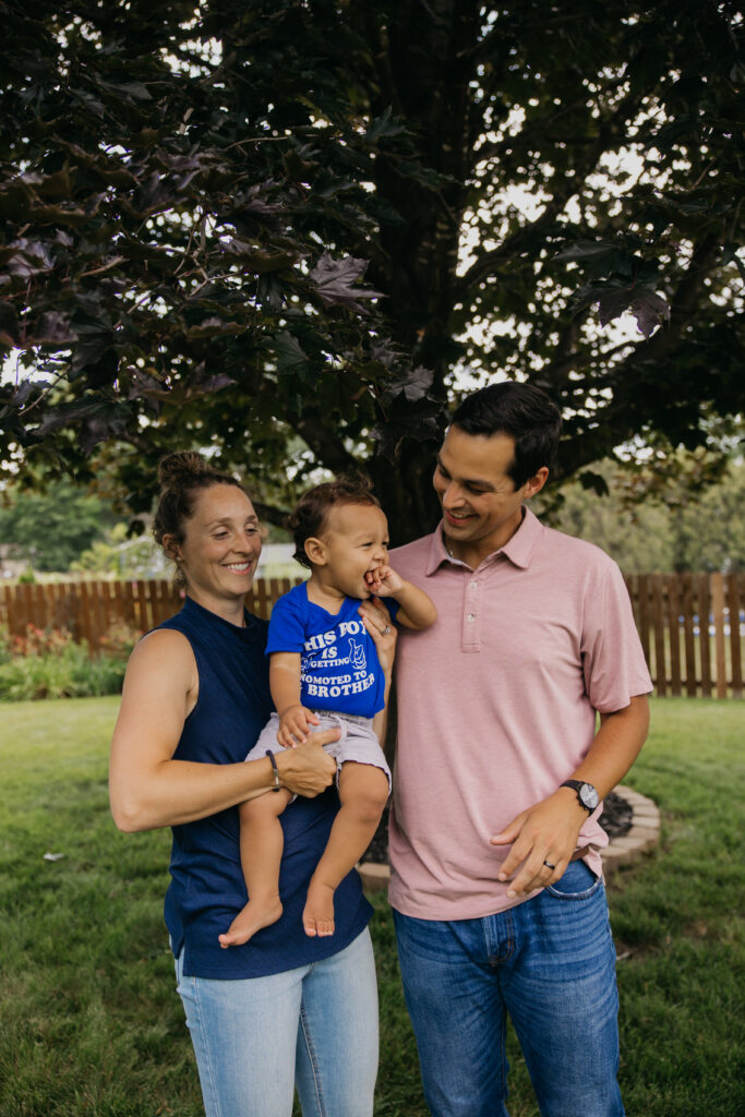 Mom holding baby in a big brother announcement shirt while dad stands close beside them under a tree