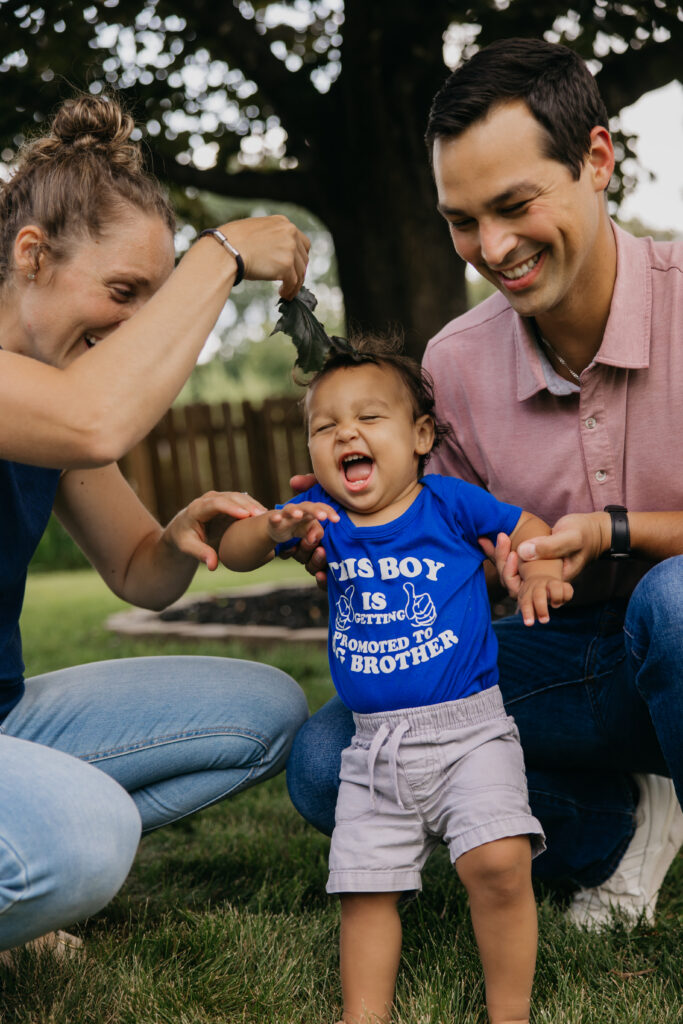 Baby laughing with arms raised as mom holds a leaf overhead and dad crouches beside him in the backyard