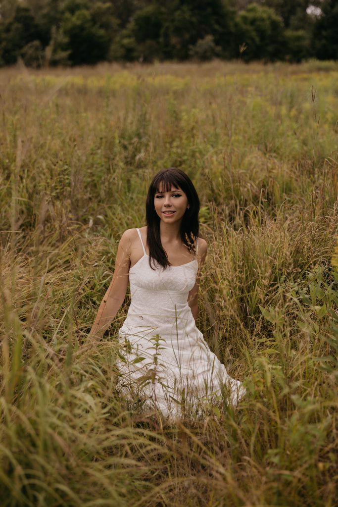 Senior with dark hair and bangs in a white dress sitting in tall golden grass