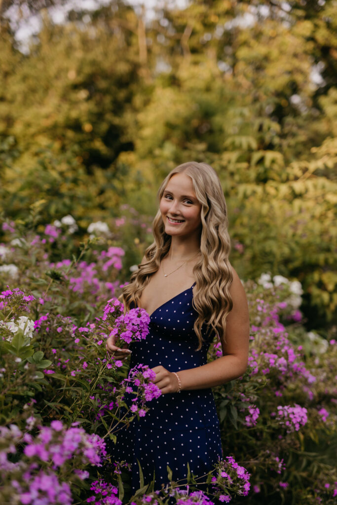 Senior in a navy polka-dot dress standing among purple phlox during Minneapolis senior photos