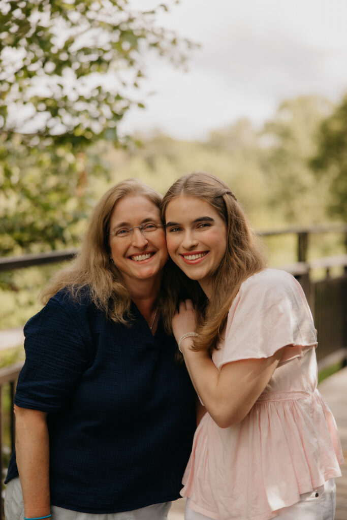 Smiling mother and senior daughter in a pink dress posing close together by a railing
