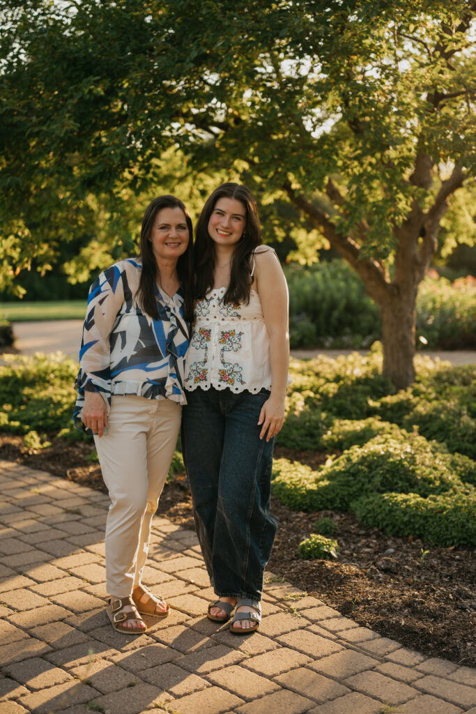 Mother and senior daughter standing together on a garden path beneath a tree at sunset