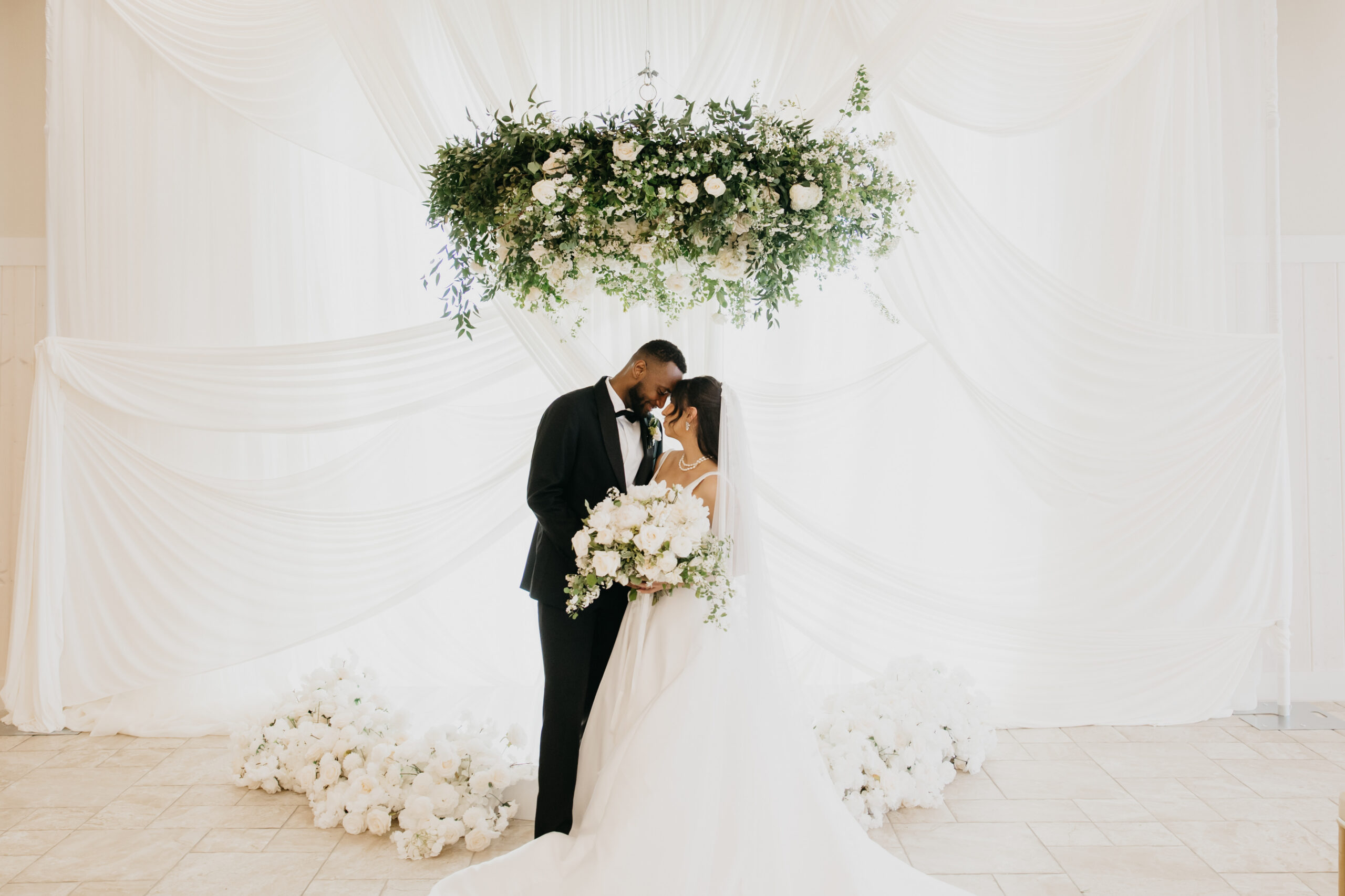 Wedding models touch foreheads under a hanging greenery and white rose installation at an Equestria West wedding styled shoot in Chaska, MN