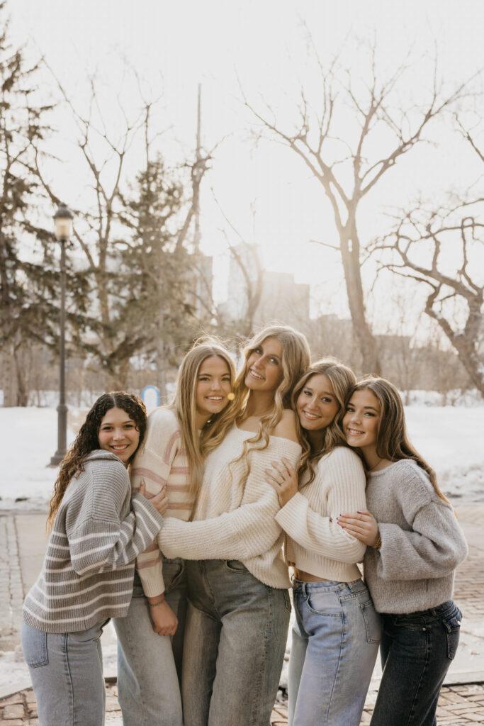 Five senior girls in cozy sweaters hugging in a snowy Minneapolis park for friend senior photos
