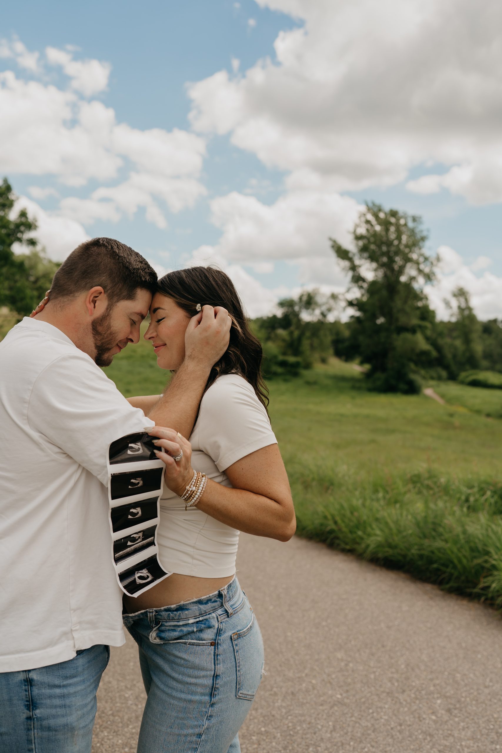 Couple touching foreheads on a paved park path while holding a strip of ultrasound photos in their pregnancy announcement photos