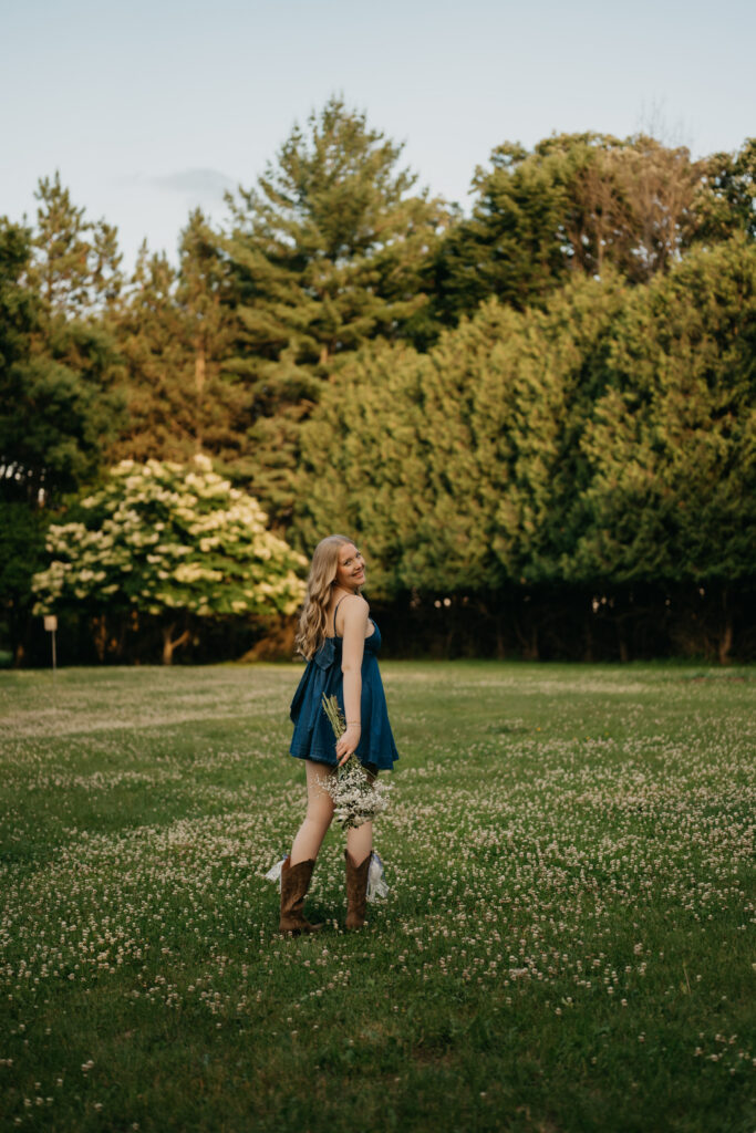 Senior girl in a denim dress and cowboy boots walking across a clover-filled lawn
