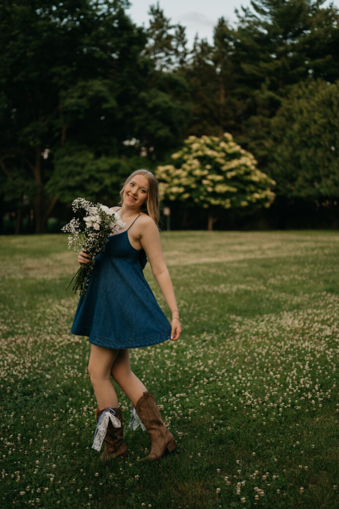 Senior in a denim dress and cowboy boots twirling with a white bouquet on a lawn