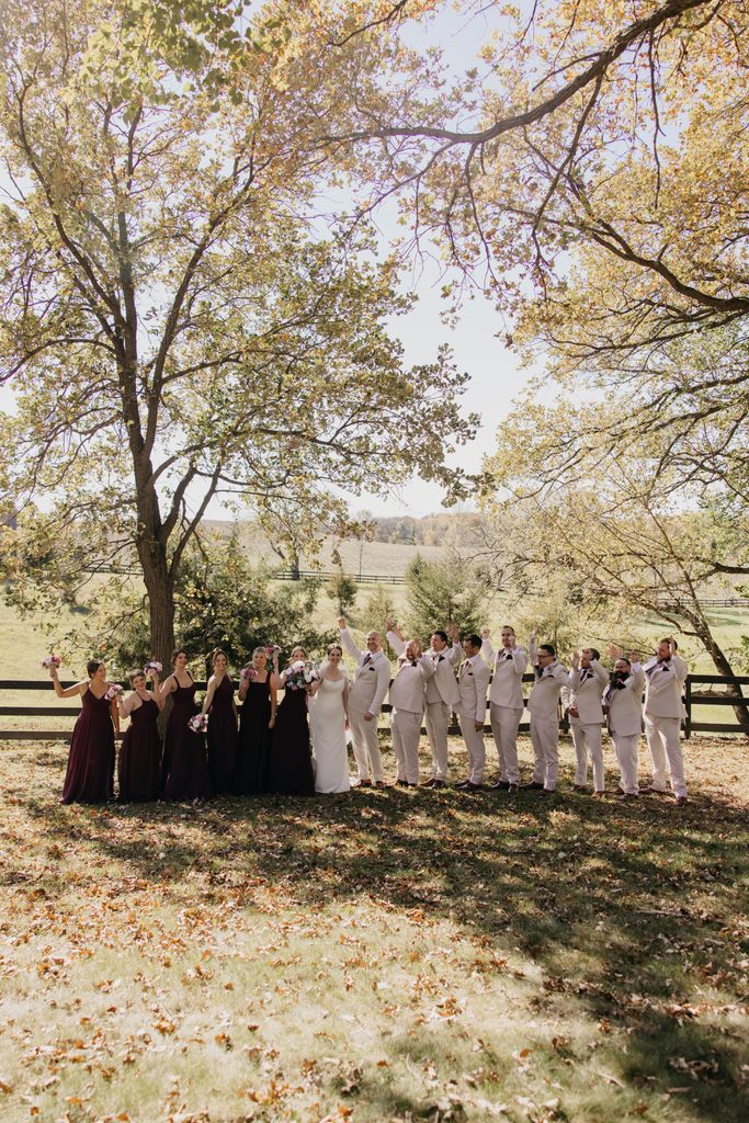 Wide view of the whole wedding party lined up beside a fence under fall trees