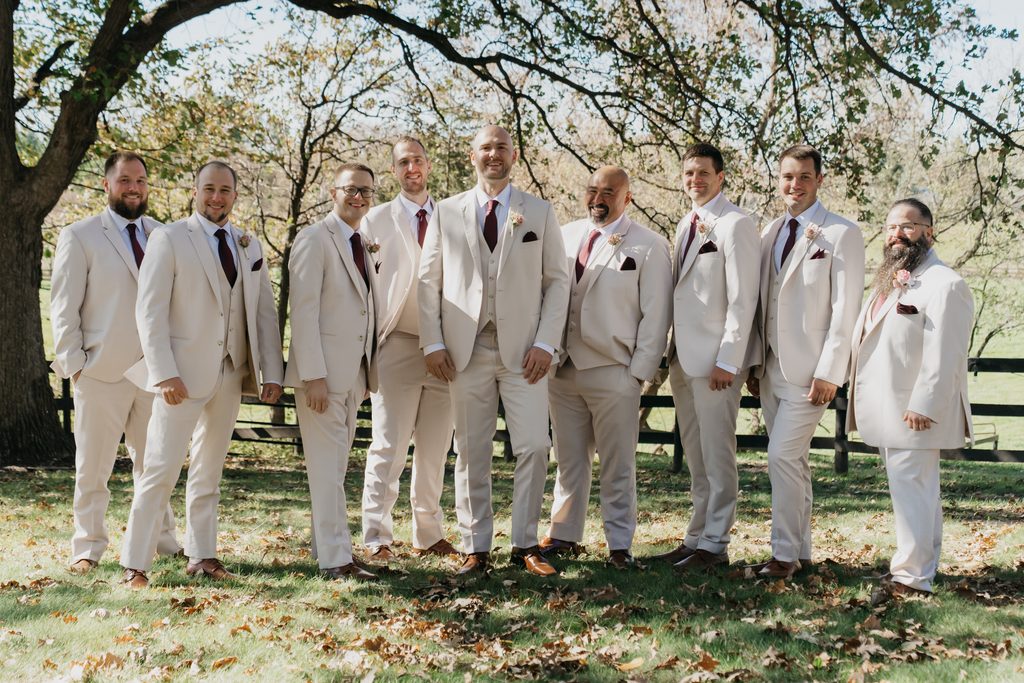 Groom and eight groomsmen in tan suits and burgundy ties standing in a row under a tree