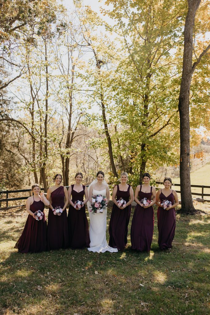 Bridesmaids and bride stand in a line holding bouquets under tall fall trees