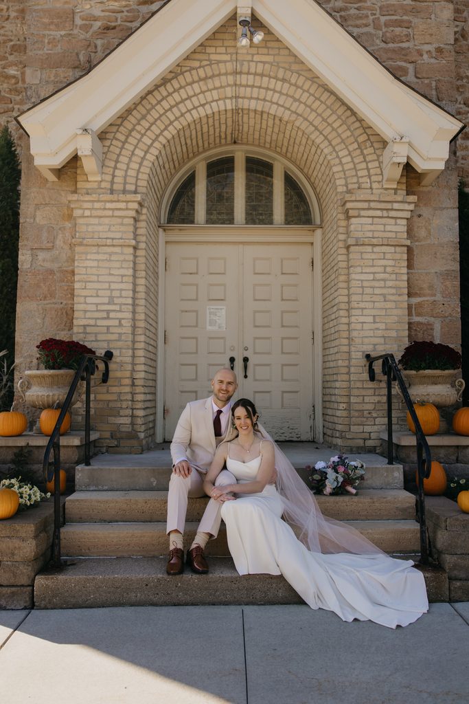 Couple sit on the church steps surrounded by pumpkins with her veil draped beside them