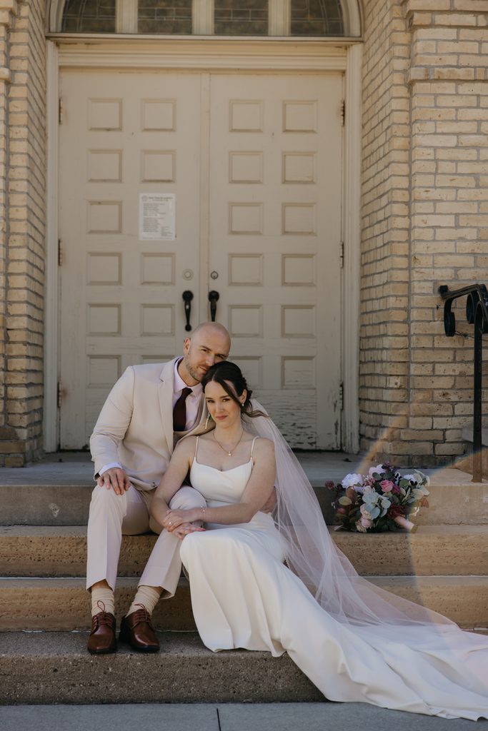 Bride and groom lean together on the church steps with her veil and bouquet