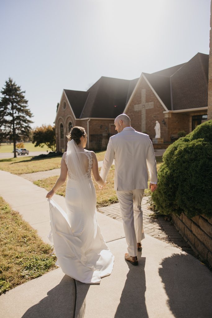 Bride lifts her long veil and train as the couple walk the church sidewalk