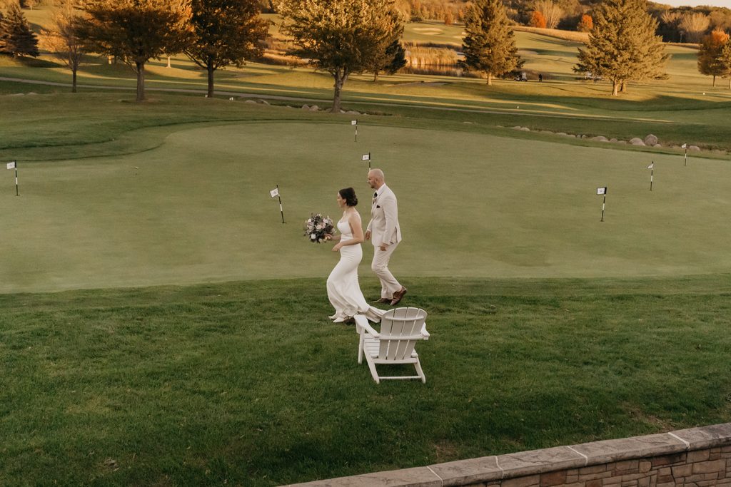 Bride carrying a bouquet and groom walk past a putting green and white Adirondack chair
