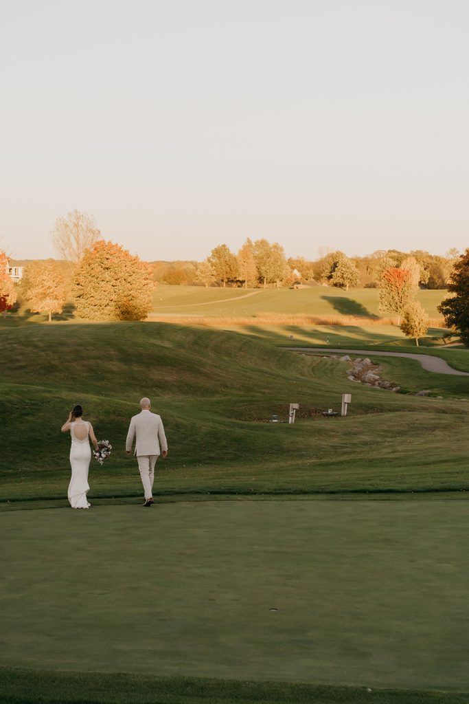 Bride carrying a bouquet and groom walk away across a golf course fairway