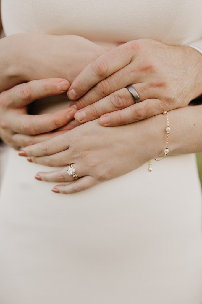 Couple’s clasped hands show her engagement ring, his wedding band, and a pearl bracelet