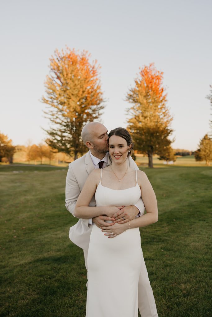 Groom kissing bride’s cheek from behind with two orange fall trees on the golf course