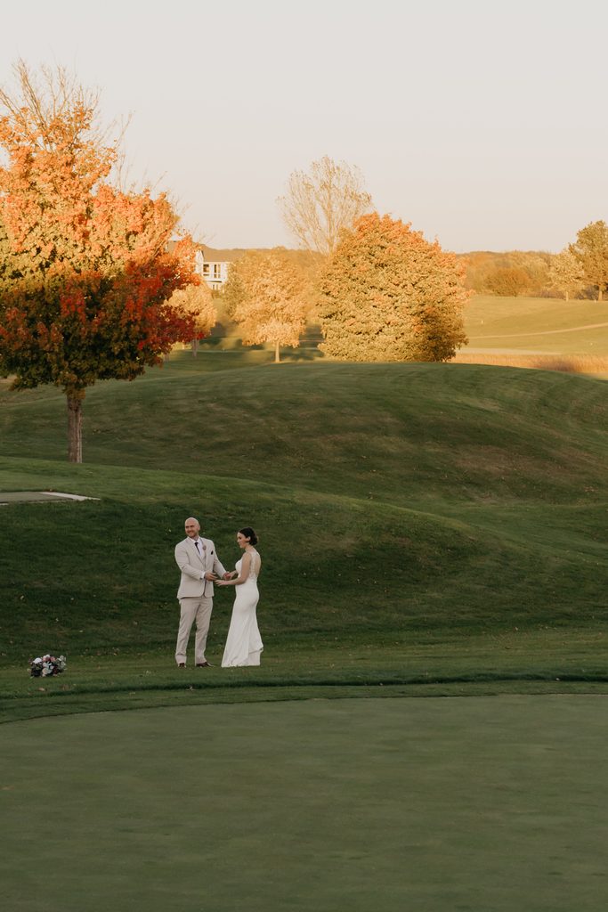 Bride and groom hold hands on a golf green with an orange maple tree nearby