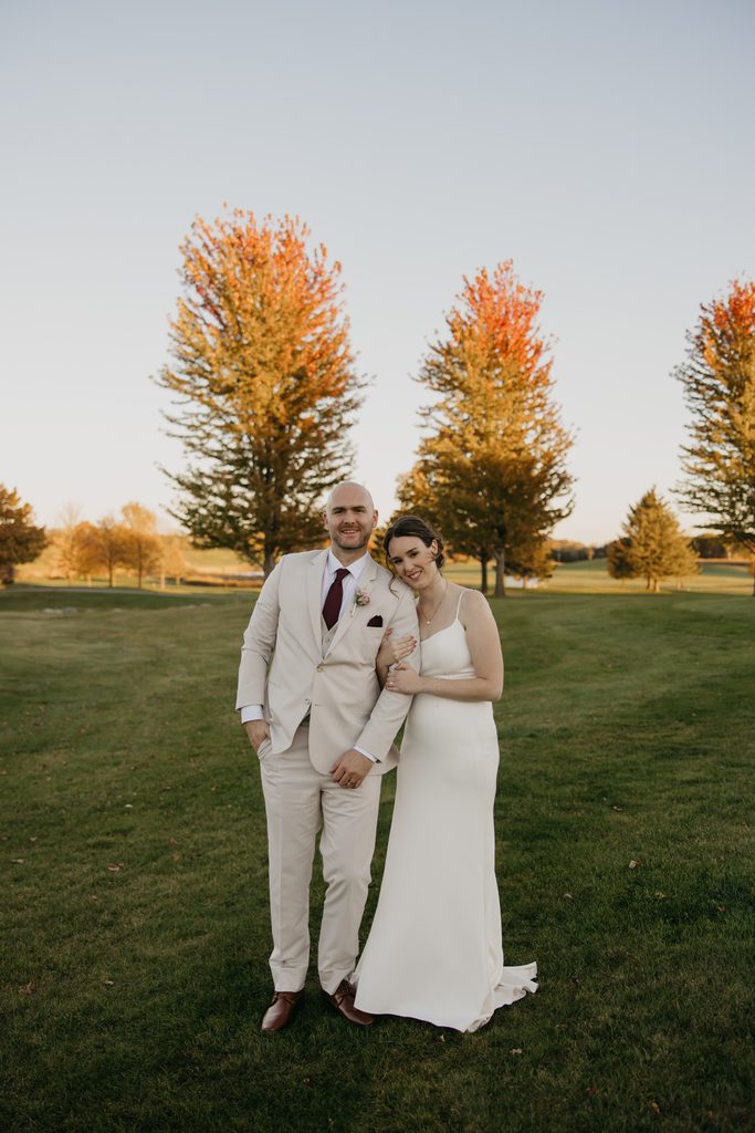 Bride and groom stand arm in arm smiling in front of three orange maple trees