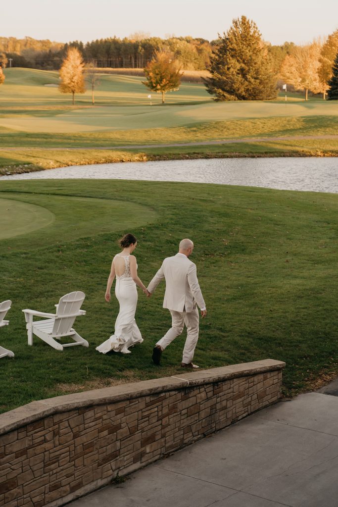 Bride and groom stroll by a golf course pond near white Adirondack chairs