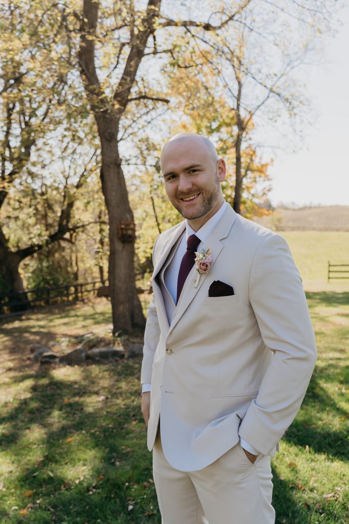 Smiling groom in a tan suit and burgundy tie stands beside a large tree