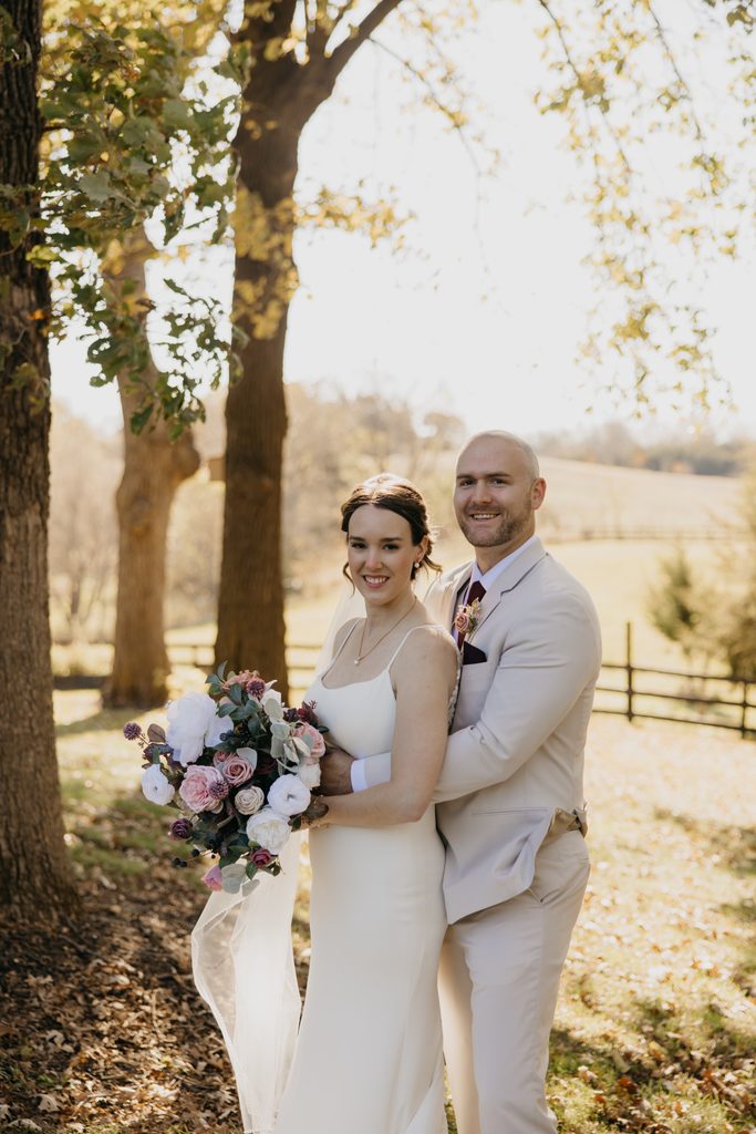 Bride with bouquet smiles as groom holds her close beside a sunlit tree