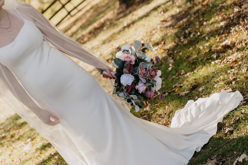 Wind lifts the bride’s gown train as she holds a bouquet on a grassy slope