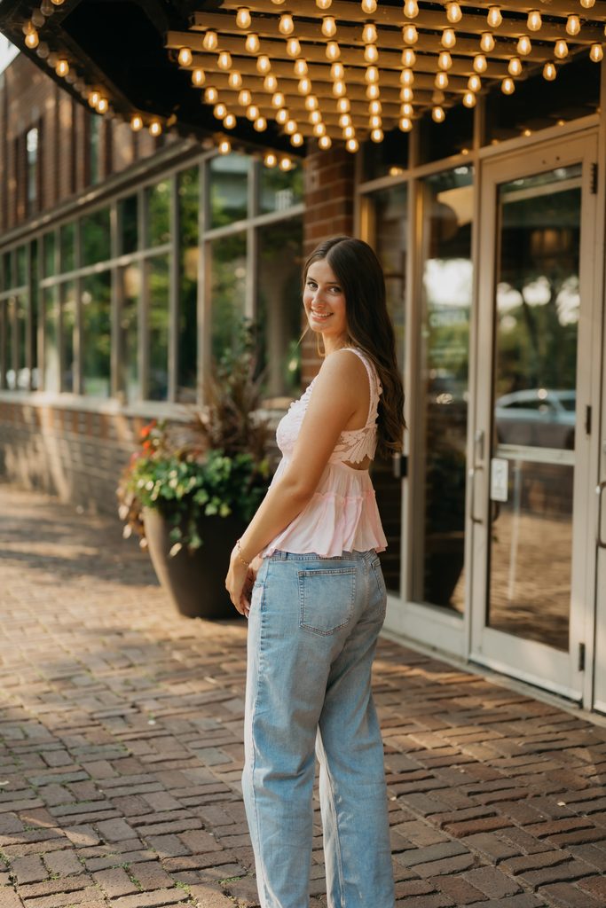 Senior in a light pink top and light-wash jeans looking over her shoulder in front of a brick restaurant with marquee lights