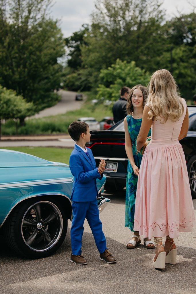 Boy in a blue suit talks with two women beside a turquoise vintage car outdoors