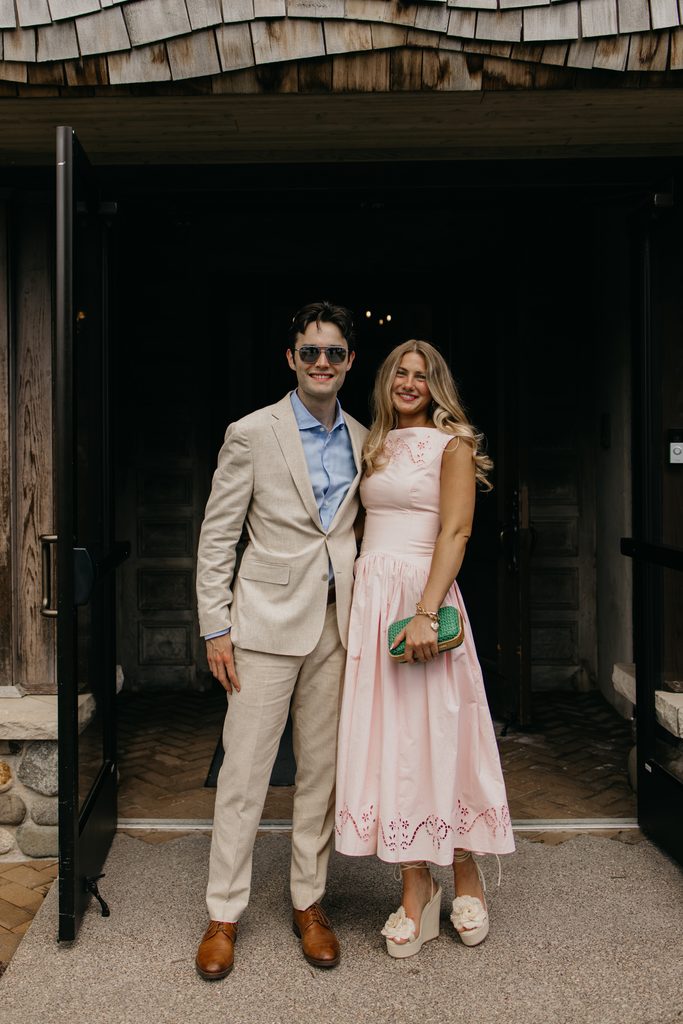 Man in sunglasses and a tan suit stands beside a woman in a pink dress in front of a dark wooden doorway