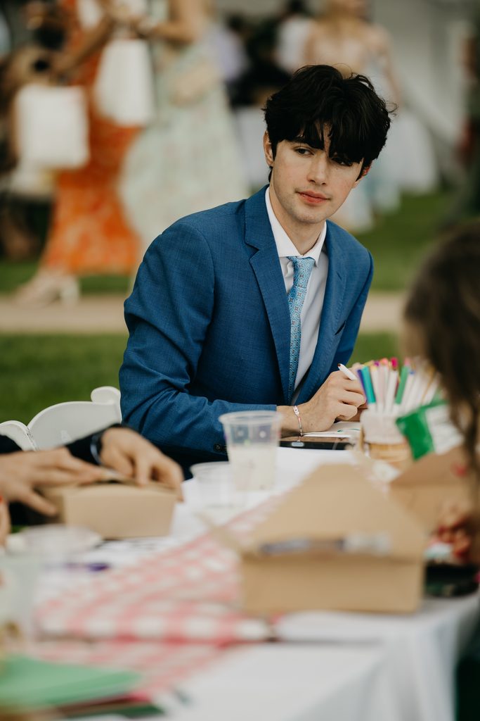Young man in a blue suit sits at a table with colored pencils and craft supplies as guests blur behind him