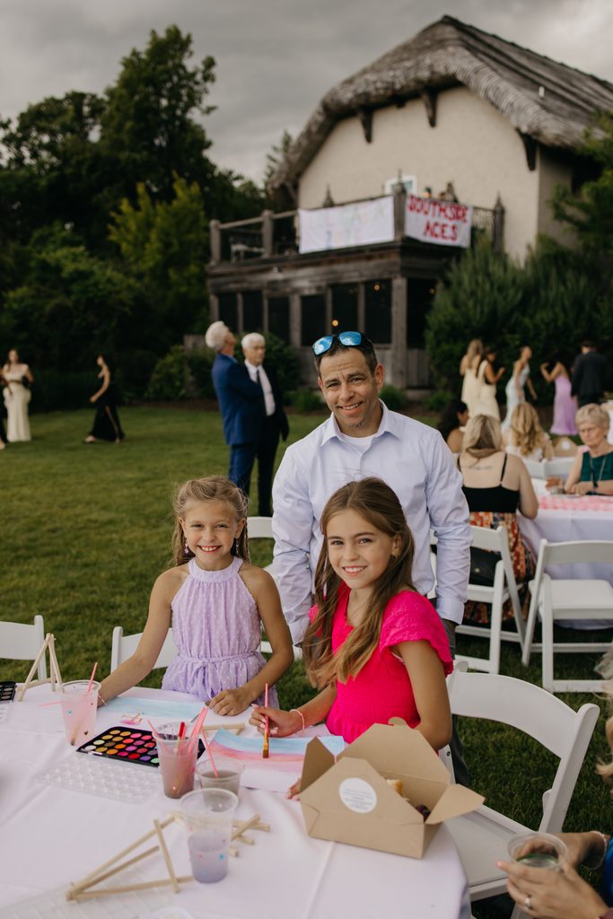 Two girls sit at a table with paint palettes while a man stands behind them, with guests and a thatched-roof building beyond