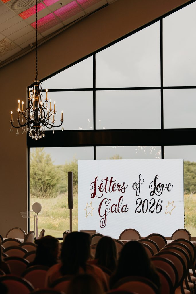 Chandelier-lit ballroom with a handwritten Letters of Love Gala 2026 sign at the front and guests seated in the foreground