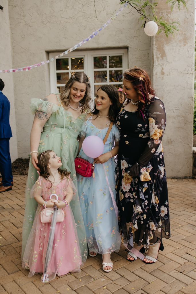 Two women and two girls in pastel and floral dresses stand together on a brick patio, one girl holding a balloon